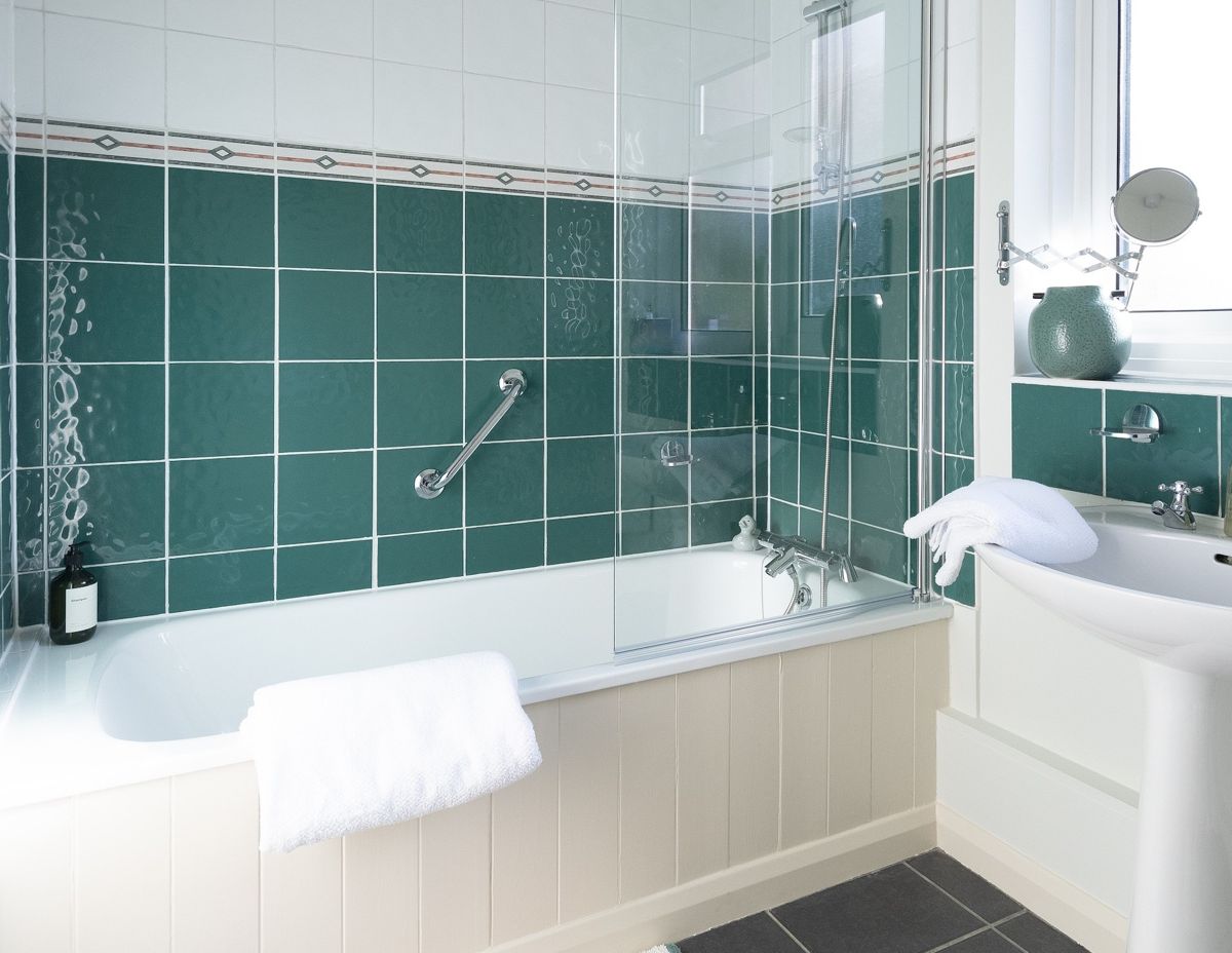 A bathroom with green tiles, a white bath with a glass screen and a shower over it, and a pedestal basin