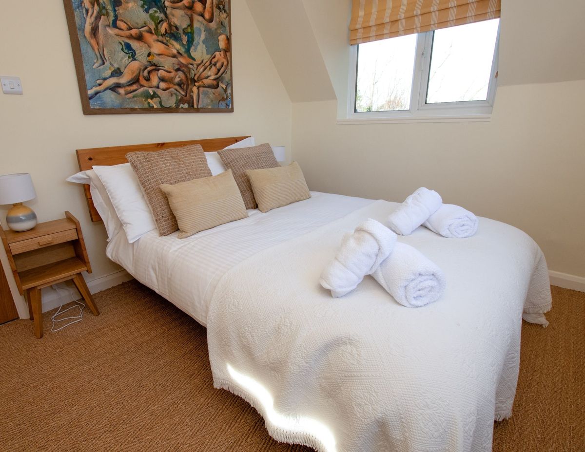 A bedroom with a double bed in white linen, cream cushions, a skylight and an abstract painting above the bed