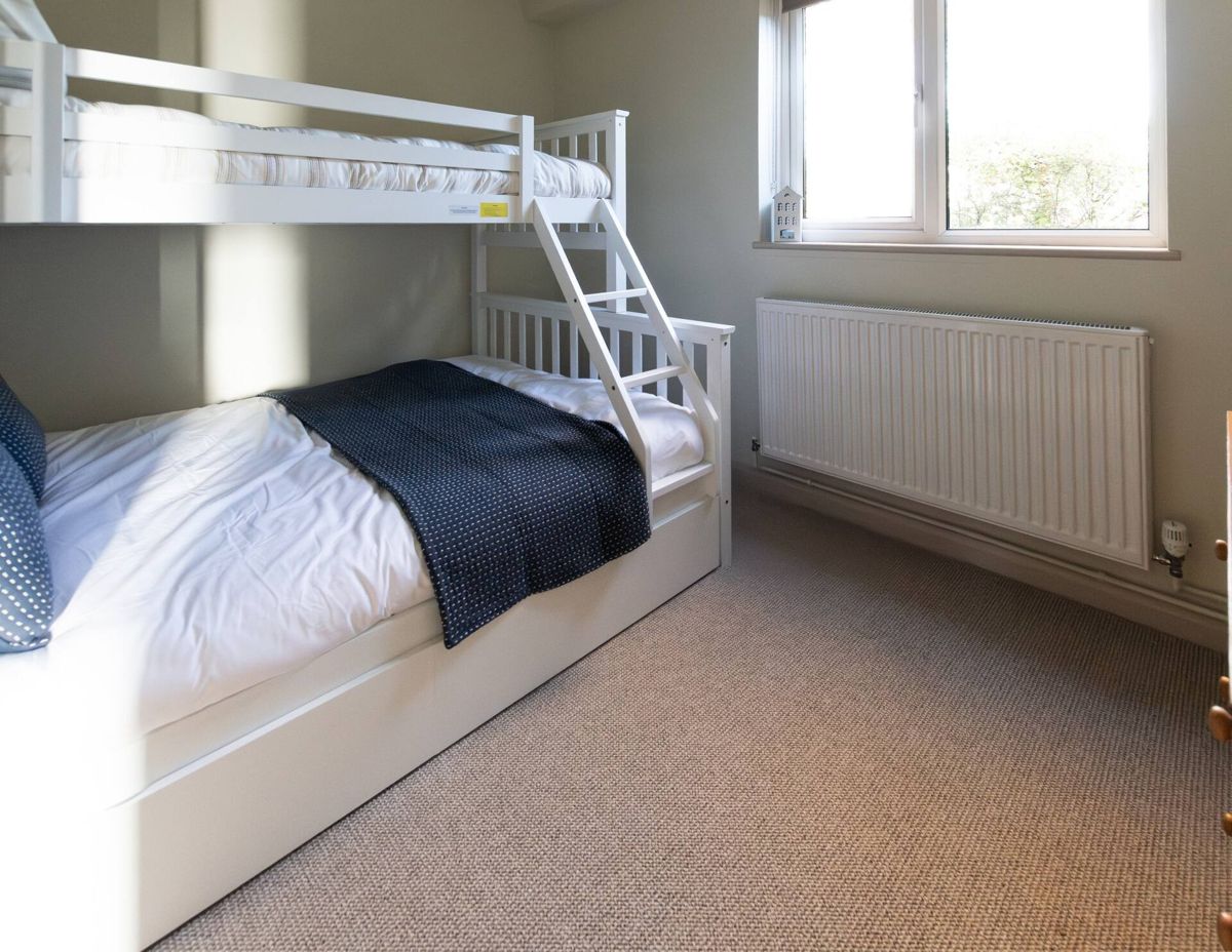A ground-floor bedroom with a white bunk bed, a double bed below with a navy throw and a single bed above, and a window