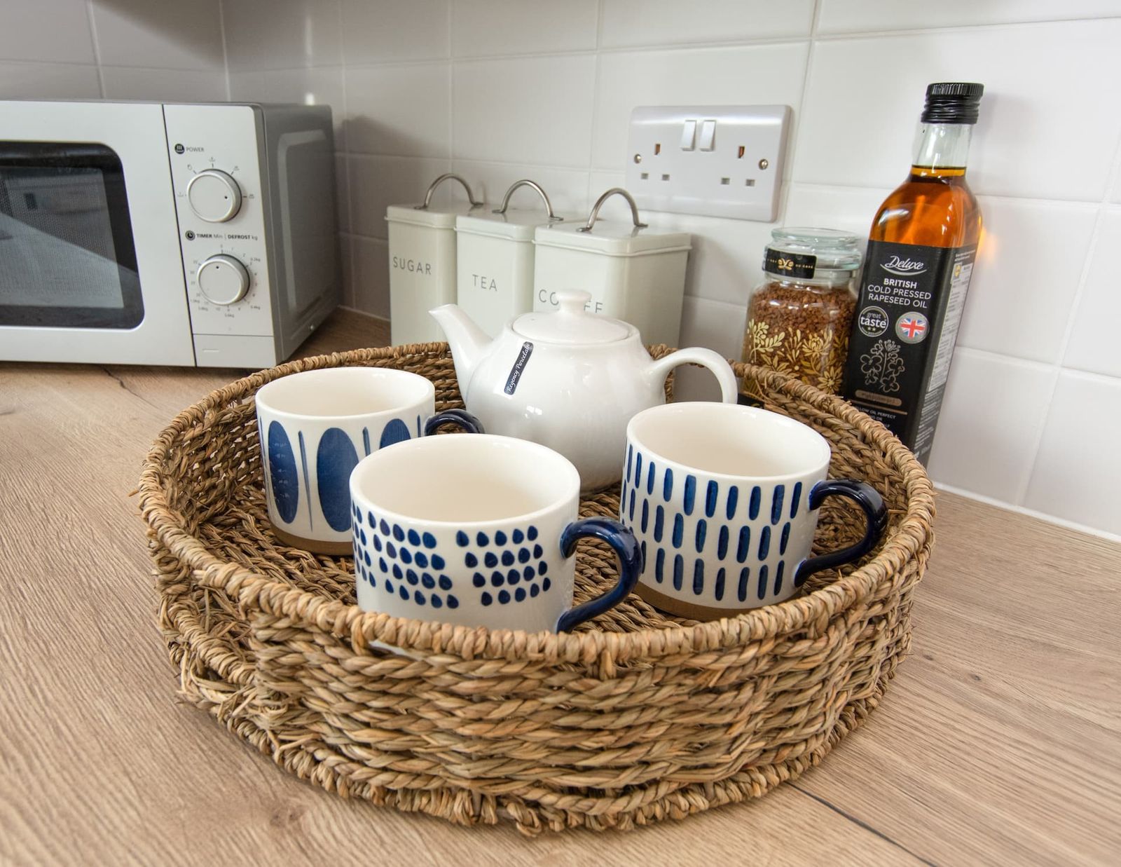 A wicker tray with a teapot, blue and white mugs and a bottle of oil on a white kitchen counter
