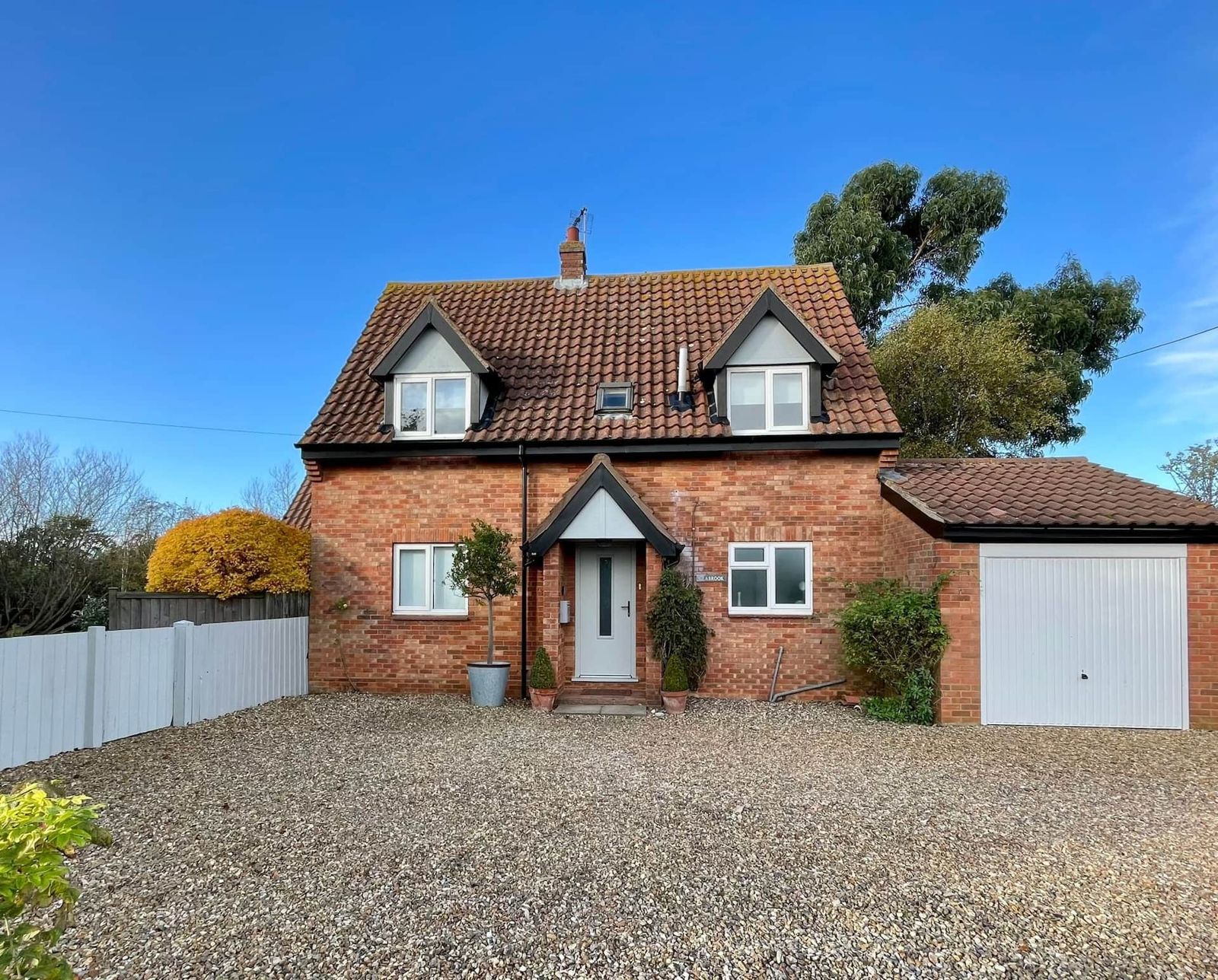 A red-brick cottage with a tiled roof, white windows and a garage, behind a wide gravel drive under a blue sky