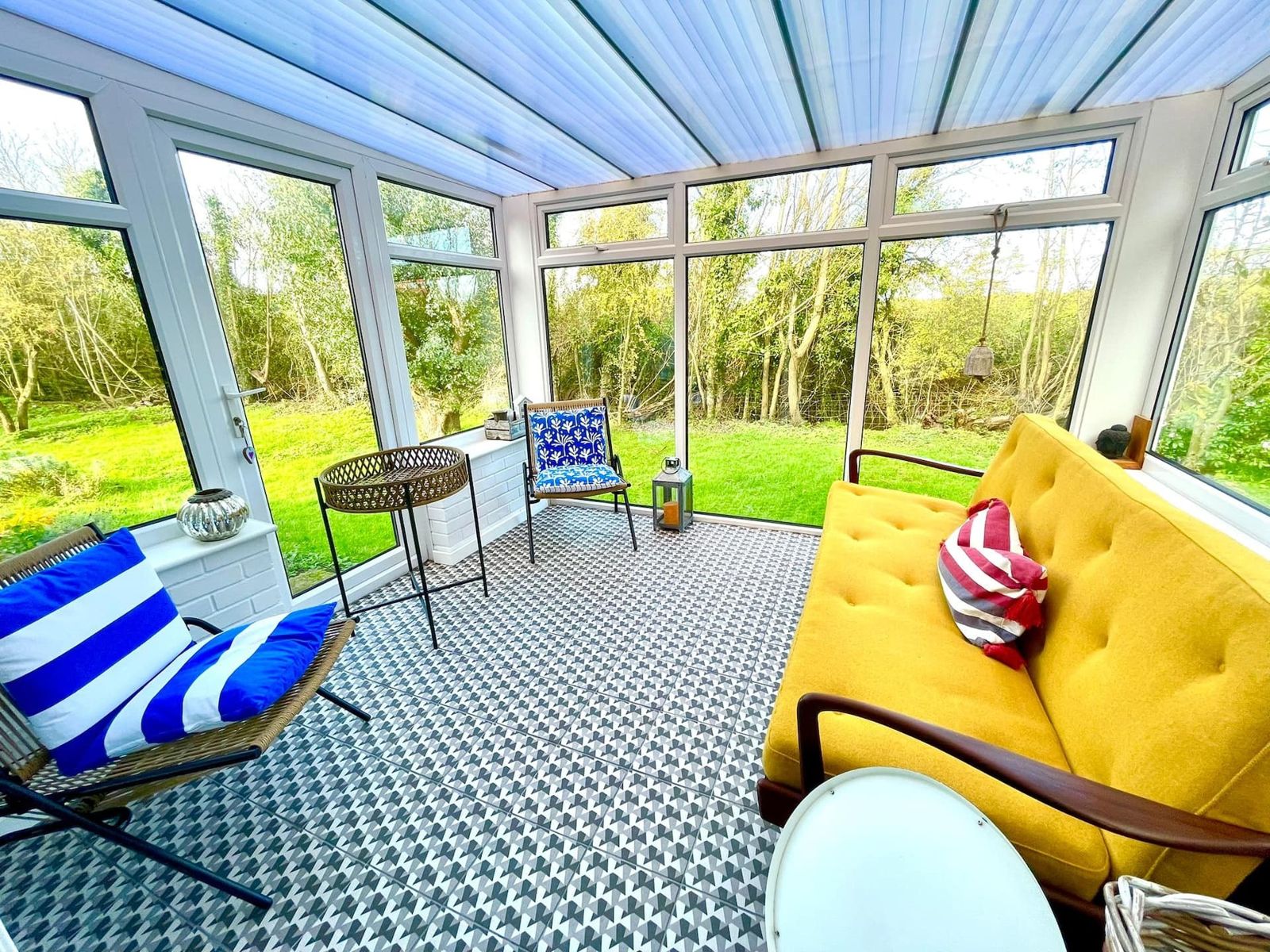 A bright conservatory with a patterned tiled floor, a yellow sofa, striped deckchairs and glass walls looking onto the garden
