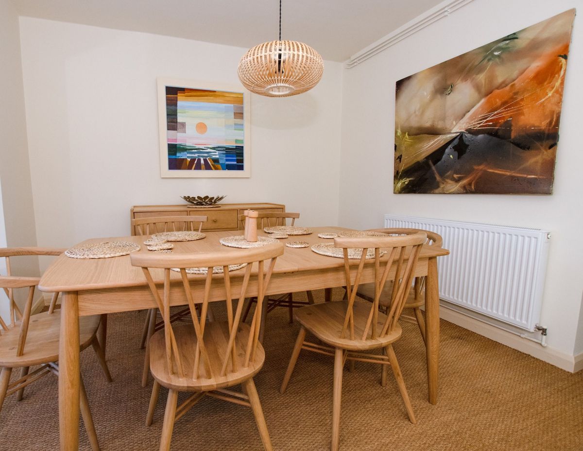 A dining room with a wooden table, spindle-back chairs, a pendant light and two colourful paintings